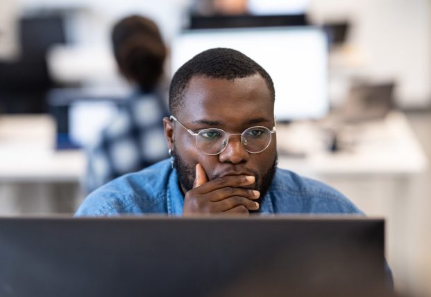 Developer with glasses looking at computer with hand over mouth concentrating on information on the screen.