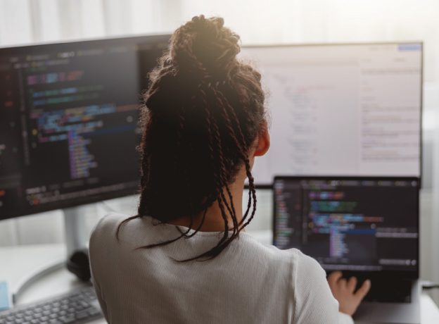 Woman working on multiple screens with code sitting in front of desk.