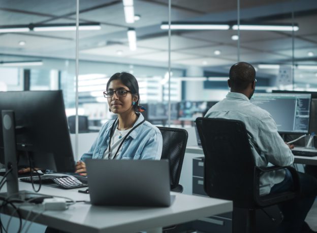 Woman developer sitting and working at desk with another co-worker sitting behind her writing code.