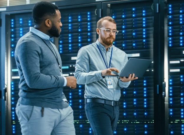 Two IT workers walking through a server room together while holding a laptop