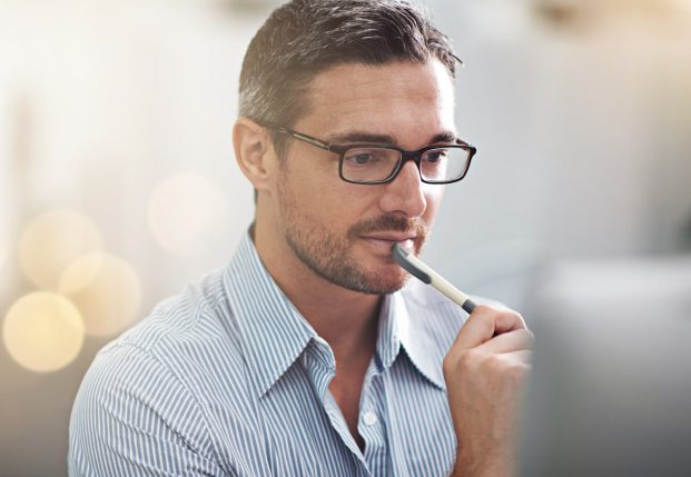 Clean cut business man with glasses holding a pen looking at a computer screen.