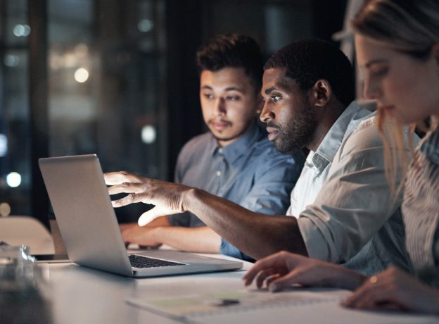 Team of developers working together at night in the office review information on a laptop.